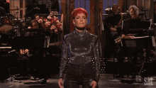a woman with red hair stands on a stage in front of an orchestra with nbc written on the bottom
