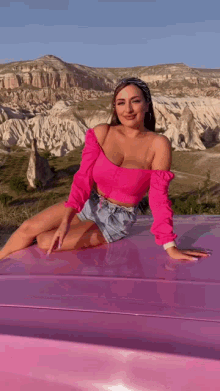 a woman in a pink top and denim shorts is sitting on the roof of a pink car .