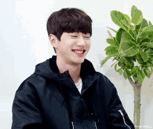 a young man in a black jacket is smiling in front of a plant