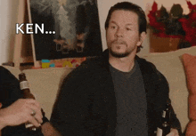 a man with a beard is sitting on a couch holding two bottles of beer and saying ken .