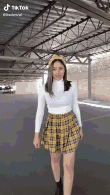 a woman wearing a white turtleneck and a yellow plaid skirt is standing in a parking garage .