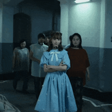 a girl in a blue dress stands with her arms crossed in a dark room