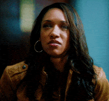 a woman wearing hoop earrings and a brown jacket looks at the camera