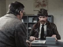 a man with a mustache sits at a desk talking to another man in a brown jacket