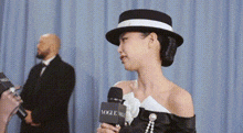 a woman in a hat is talking into a microphone while standing in front of a blue curtain .