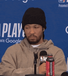 a man wearing a beanie stands in front of a microphone holding a bottle of bodyarmor