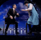 a man in a suit and tie is sitting on a chair while a woman in a blue dress is dancing on a stage