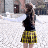 a woman in a plaid skirt is standing on a cobblestone street with her arms outstretched