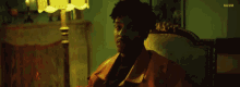 a man in an orange jacket is sitting in a chair in front of a lamp in a dark room .