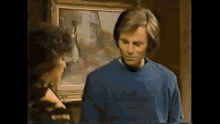 a man in a blue sweater is standing in front of a painting .
