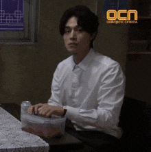 a man in a white shirt and tie sits at a table in front of a sign that says ocn