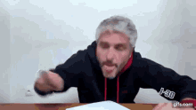 a man with gray hair and a beard is sitting at a table holding a piece of paper and pointing at the camera .
