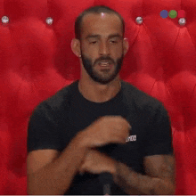 a man with a beard is sitting in front of a red couch and holding his hands together .