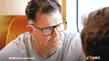 a man with glasses and a beard looks at another man with the nbc destination x written on the bottom
