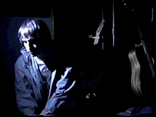 a man in a blue jacket stands in a dark room with a guitar in the background