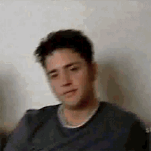 a man in a gray shirt is sitting in front of a white wall and looking at the camera .