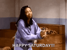 a woman in a purple sweatshirt is dancing in a room with the words happy saturday written above her .