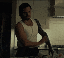 a man in a white tank top is standing in a kitchen holding his arm .