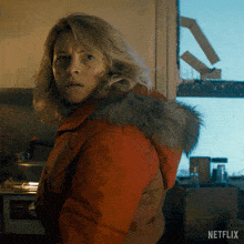a woman in an orange jacket with a fur hood is standing in front of a netflix logo