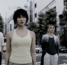 a woman in a white tank top stands in front of a man in a gray cardigan