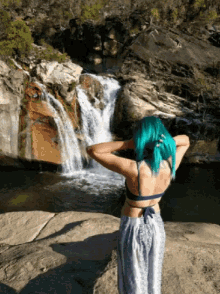 a woman with blue hair is standing on a rock near a waterfall