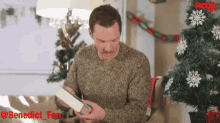 a man in a sweater is reading a book in front of a christmas tree with hook written on the bottom