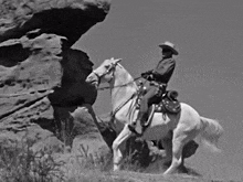 a man in a cowboy hat is riding a white horse on top of a rocky hillside .