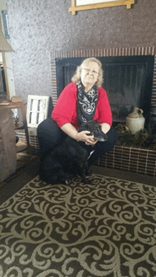 a woman in a red shirt is kneeling down holding a black dog in front of a fireplace