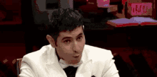 a man in a white suit and bow tie is sitting at a table with his mouth open .