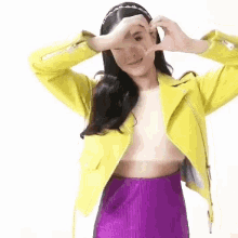a woman in a yellow jacket and purple skirt is making a heart shape with her hands .