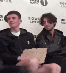 two men are sitting in front of a big web live sign