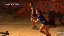 two female athletes are running on a sandy field with a scoreboard in the background that says titanes 8 5 heroes