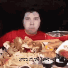 a man in a red shirt is sitting at a table with a bunch of food and the words in hebrew