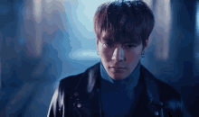 a young man wearing a black leather jacket and a blue turtleneck is standing in a dark room .