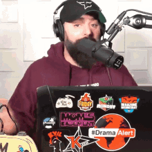 a man wearing headphones and a hat stands in front of a microphone behind a laptop with stickers on it