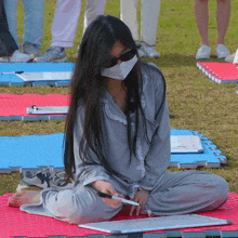 a woman wearing a mask and sunglasses sits on a mat holding a marker