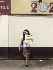 a girl is holding two certificates in front of a sign that says class of 2020