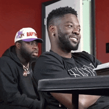 two men are sitting at a table one wearing a raptors hat