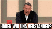 a man in a judge 's robe is sitting in front of a microphone with the words haben wir uns verstanden written above him