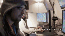 a man in a hooded jacket sits at a desk with a computer monitor and the words observers room below him