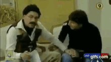 a man with a mustache is talking to another man while sitting on a couch ..