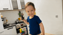 a little boy in a blue shirt is standing in a kitchen holding a spoon and smiling