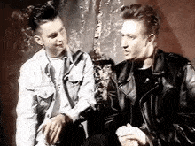 two men are sitting next to each other in a room and talking . one of the men is wearing a leather jacket .