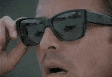 a man wearing sunglasses is taking off his sunglasses and looking at the camera .