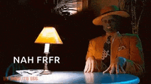 a man with a mask on his face is sitting at a table with a lamp and says nah frfr .