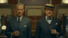 two men in suits and hats are standing next to each other and one of them is holding a packet of seagrams