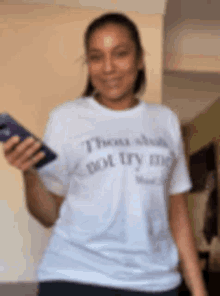 a woman wearing a white t-shirt that says `` those who do not try me '' is holding a cell phone .