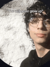 a young man with curly hair and glasses says " you will never glow up " in front of a white wall