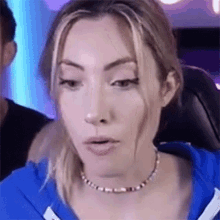 a woman wearing a necklace and a blue shirt is sitting in a chair and making a funny face .