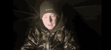 a man wearing a camouflage jacket and a beanie is sitting under an umbrella in the dark .